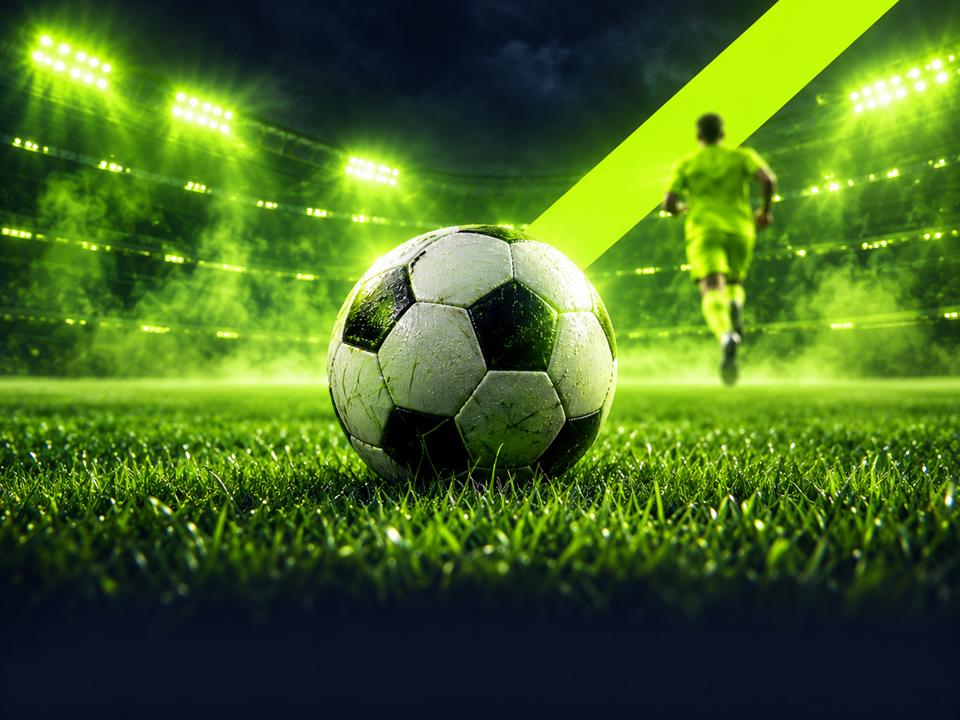 Fotball på gresset foran et opplyst stadion med limegrønne lyskastere